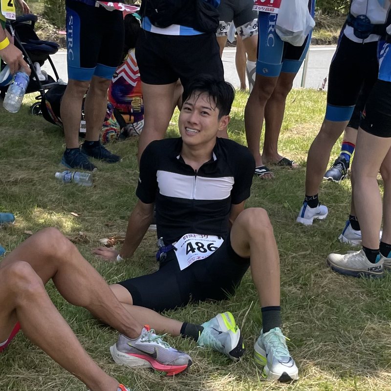 Jin in race bib 486 sitting on the grass among other athletes after the Lake Haruna triathlon